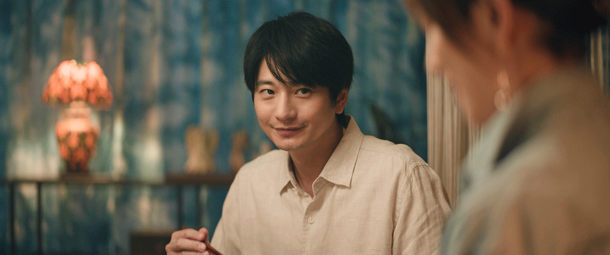 青いカーテンのかかった室内で、白いシャツを着た男性がほほえんで右の方を見ている。右側手前にいる人物はピントがぼやけている。男性は手に箸を持っている。