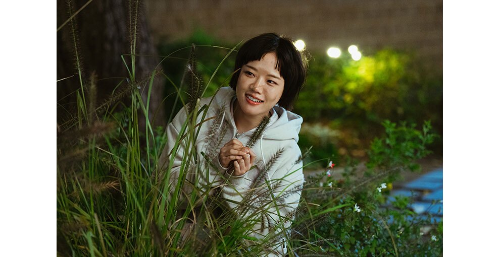 薄暗い屋内、草むらにひそみ、生い茂った雑草の影から左側をのぞき見る少女。