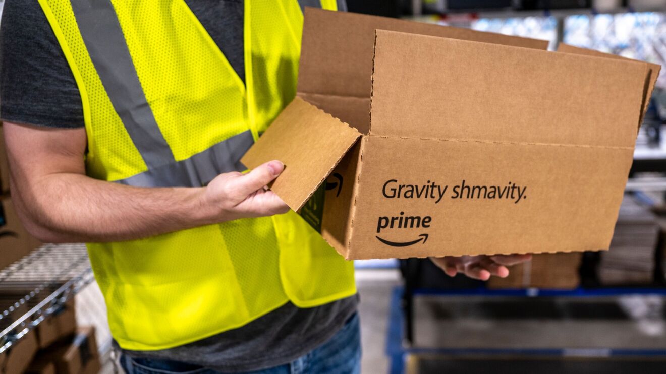 An Amazon employee holds a delivery box for a product to be delivered by drone. The text on the box says "gravity shmavity" with the Prime logo on it.