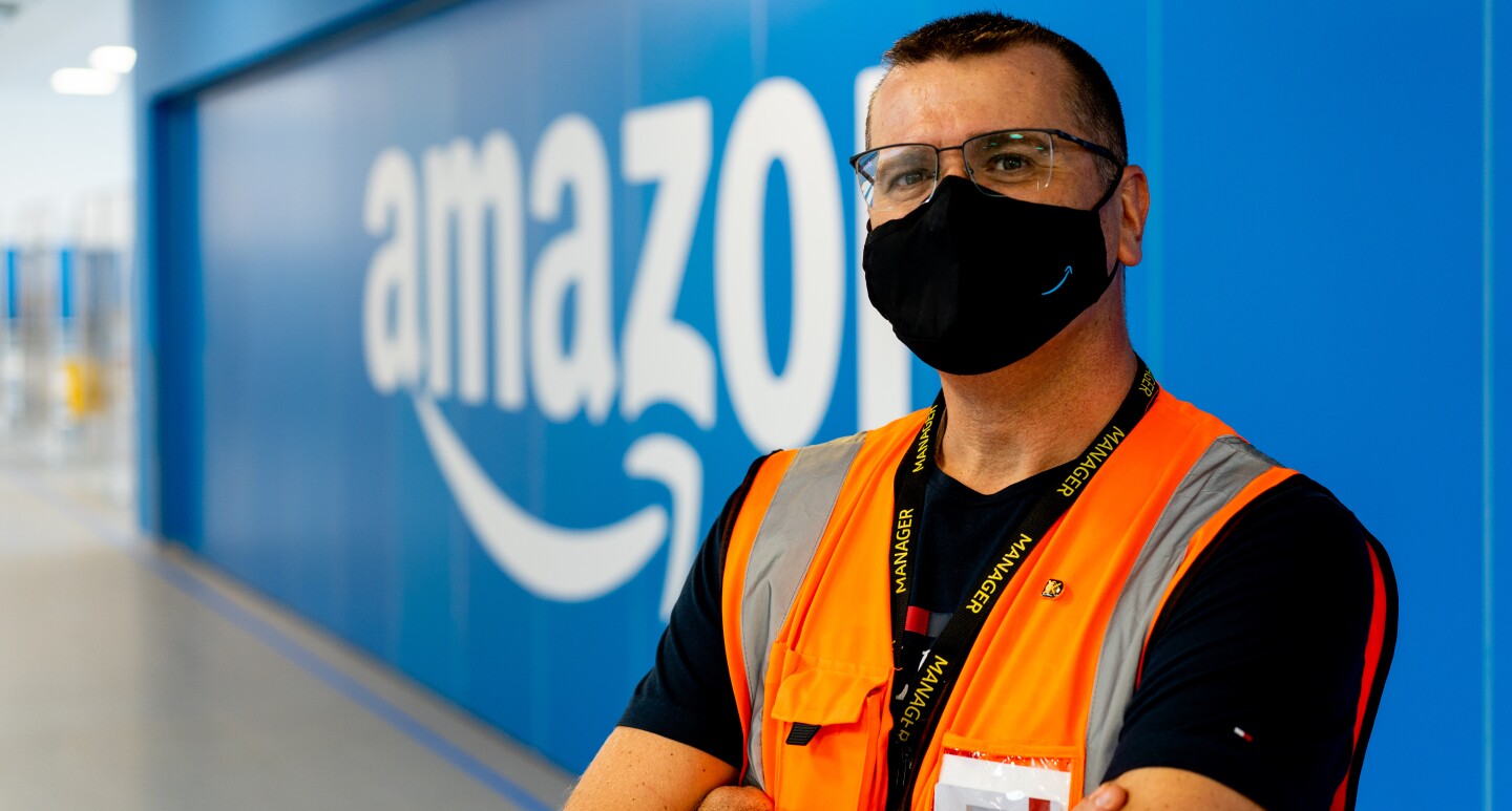 Carlos Pallarès. mánager de operaciones en Illescas. De fondo una pared de color azul con el logo de Amazon en blanco. Con los brazos cruzados y mirando a cámara está Carlos. Lleva una camiseta negra y un chaleco naranja. Tiene el pelo corto, lleva una mascarilla negra de Amazon y unas gafas negras.