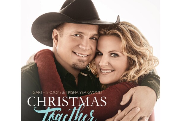 Garth Brooks and Trisha Yearwood face the camera, smiling, as they share an embrace. Garth wears a black cowboy hat and button down. Trisha wears her hair in loose curls and a burgundy long-sleeve top.