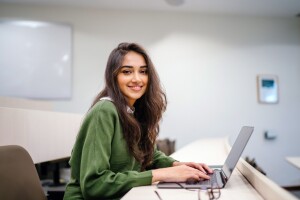 A young woman wearing a green sweatshirt smiles at the camera while studying on her laptop.