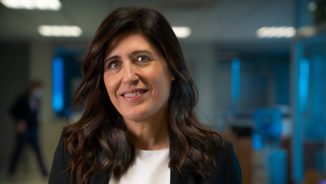 Ruth Alonso con una americana negra y un jersey gris. Sonriente mira a cámara sin mascarilla. Tiene el pelo negro por debajo de los hombros. De fondo desenfocado lo que podría ser una oficina. 