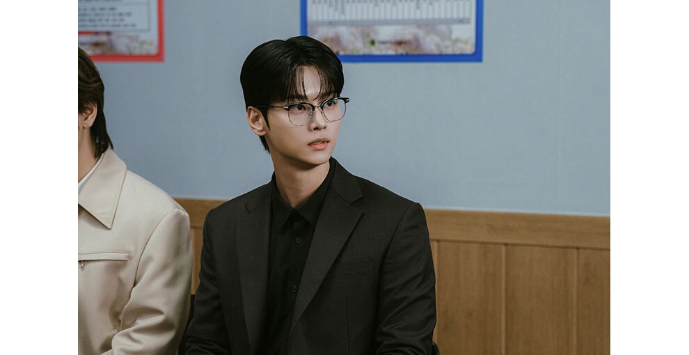 室内で着席し、右側の方に顔を向ける眼鏡の男性。左側にはもう1人並んでいる。背後は、上が白く下が木製の壁。