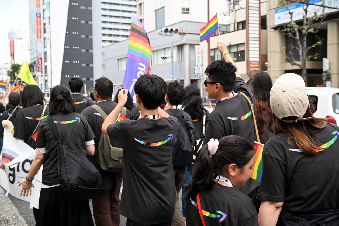 パレードをしている人々の後ろ姿。彼らはレインボー色のアマゾンロゴが印刷されたTシャツを着ている。サングラスをかけている男性がレインボーフラッグを振っている。