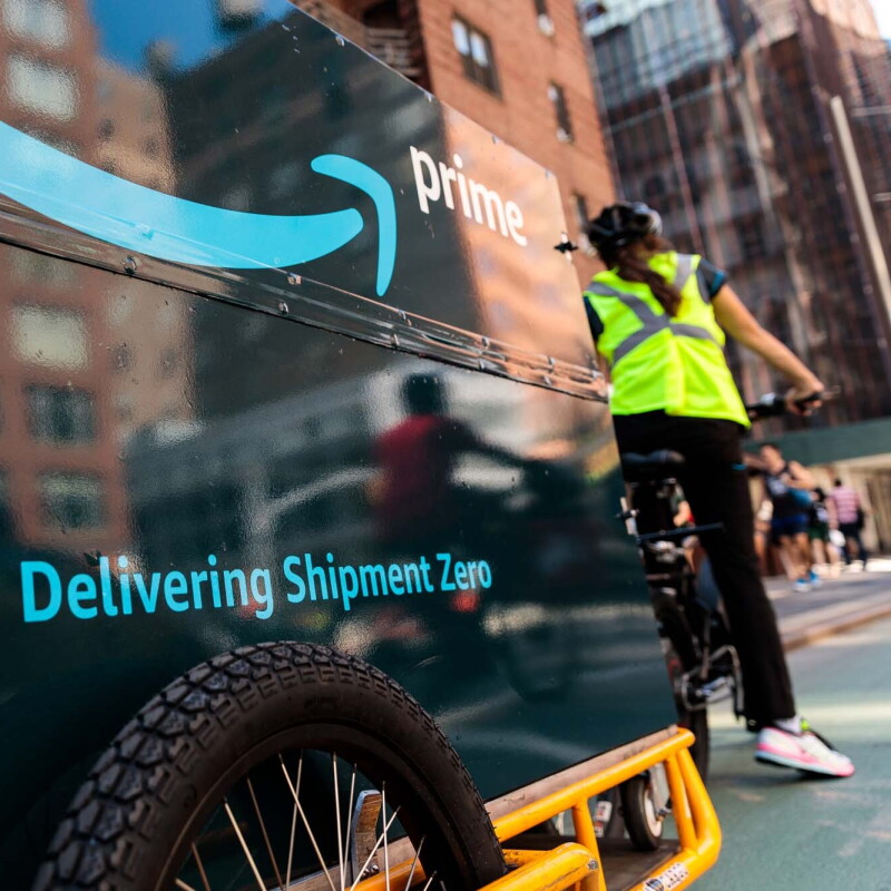 An Amazon delivery person riding a bicycle, with a bike trailer behind it. The trailer has "Delivering Shipment Zero" printed on it. 