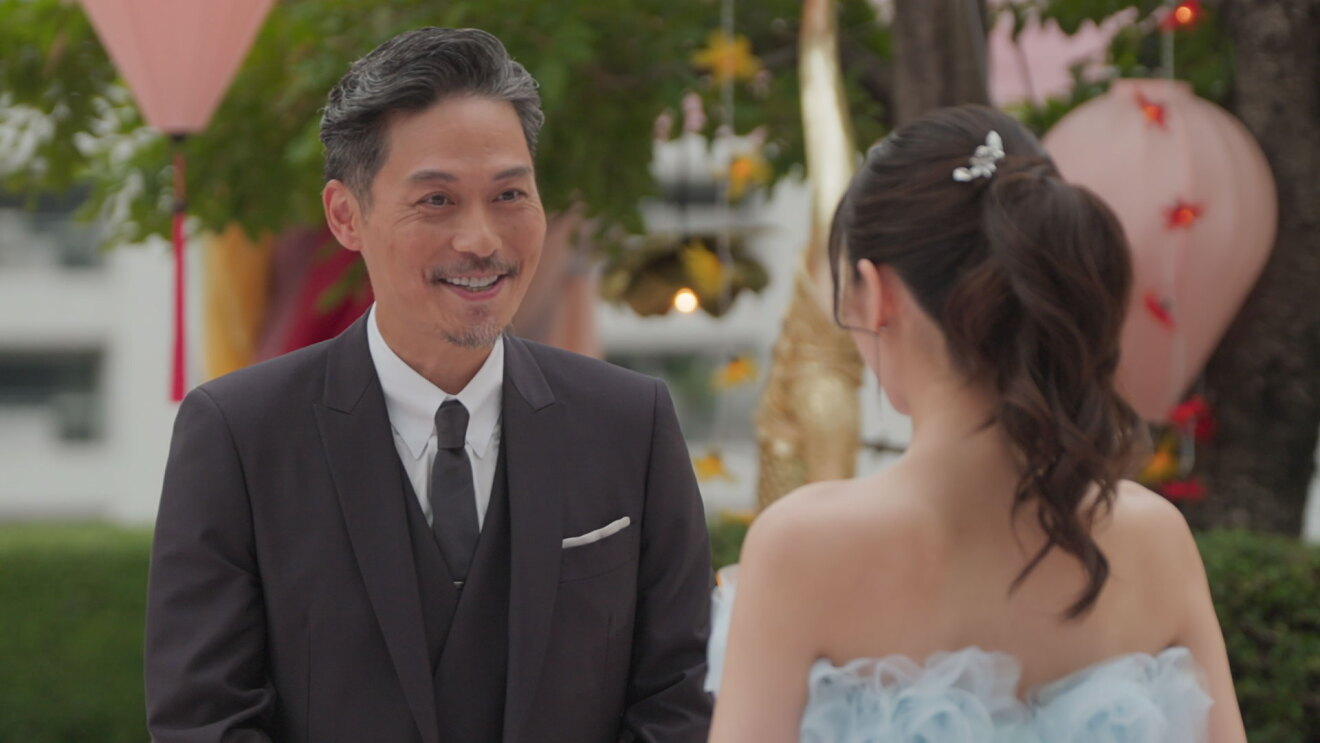 屋外、植木や木を背景に三つ揃いスーツの男性が笑顔でドレスの女性と対峙している。右側の女性は後ろ姿だけ見える。