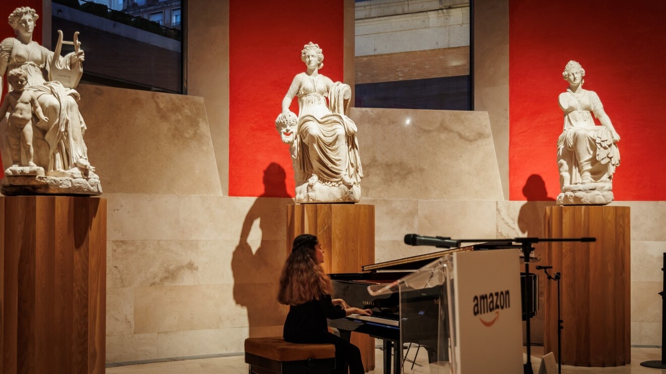 Piano Yamaha de cola negro con la tapa abierta. Plano general de Cristina Hernán Sanz, pianista residente del Museo del Prado tocando el piano en una sala ovalada del museo con tres escultura clásicas de mármol qu están colocadas en un pedestal de color madera. Detrás de cada escultura la pared es de color rojo. Hay dos grandes ventanales.