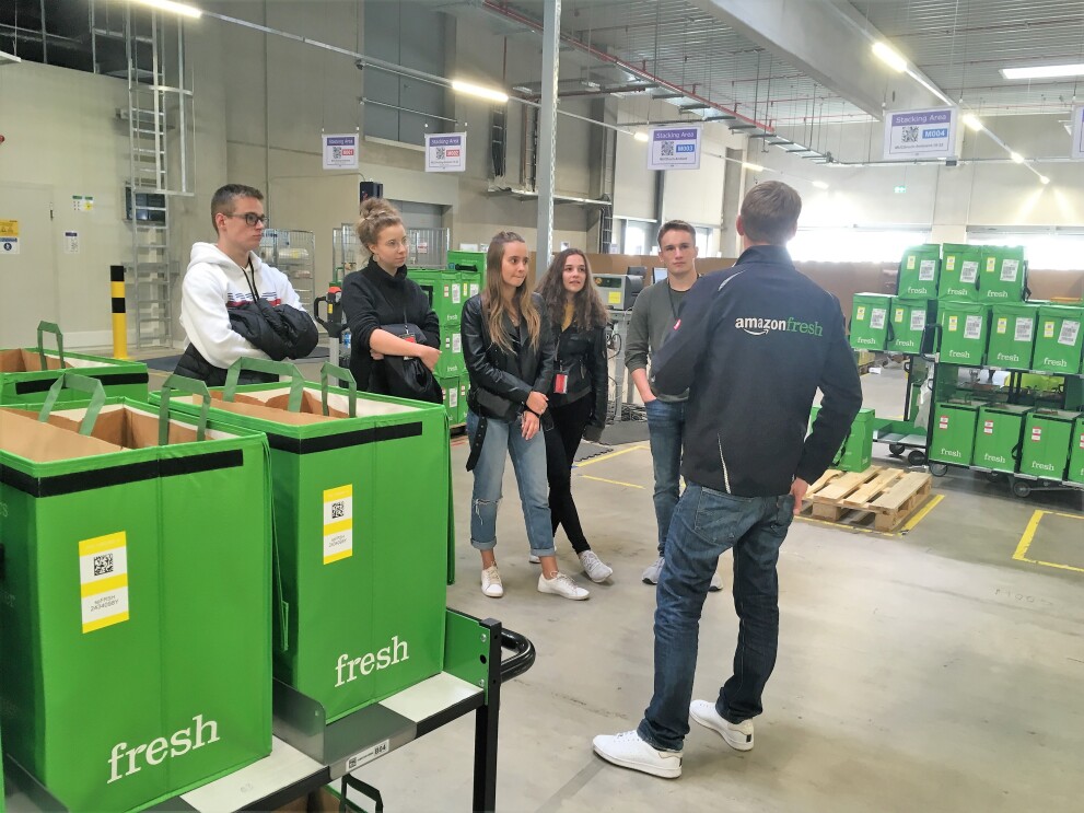 Ein Fresh Mitarbeiter ist von hinten in einem Depot zu sehen. Überall stehen grüne Boxen, in denen sich die Bestellungen für die Kunden befinden. Er trägt eine schwarze Jacke mit Aufdruck "Amazon Fresh". Vor ihm stehen die fünf jugendlichen Gewinner von "Jugend gründet". 