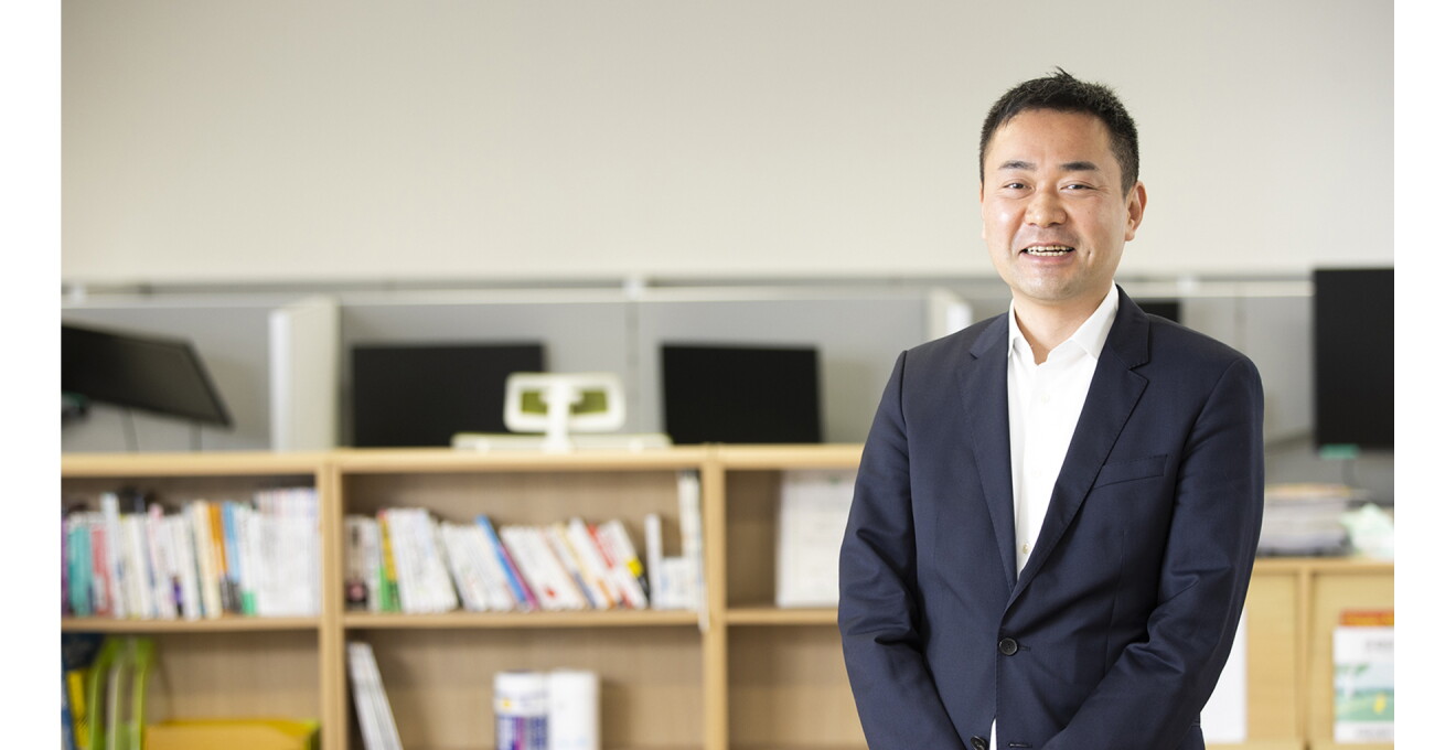 たくさんのモニターや本を背景にした谷川さんの写真