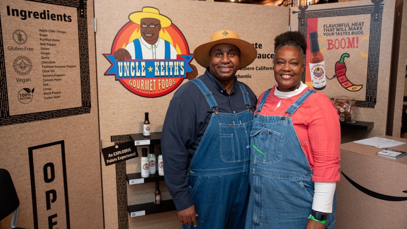 A man and a woman wearing overalls smile in front of sign that reads, "Uncle Keith's Gourmet Foods."