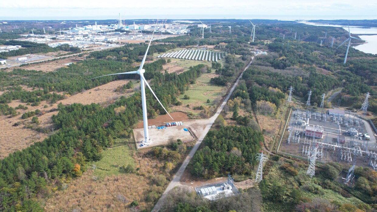 木々や畑の中に風力発電用の風車が複数立っている様子を写した空撮写真。右側には発電所の施設。