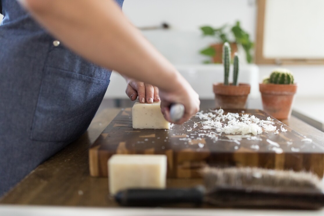 A woman hand-cuts a block of soap on a large, wooden cutting board. In front of the soap block are small bits of shavings from soap cuttings.