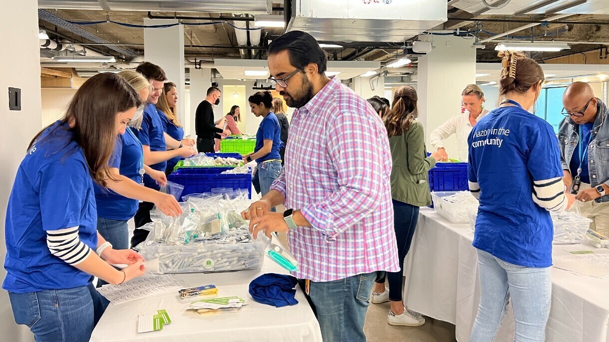 Amazon employees work together to pack hygiene kits while standing at tables.