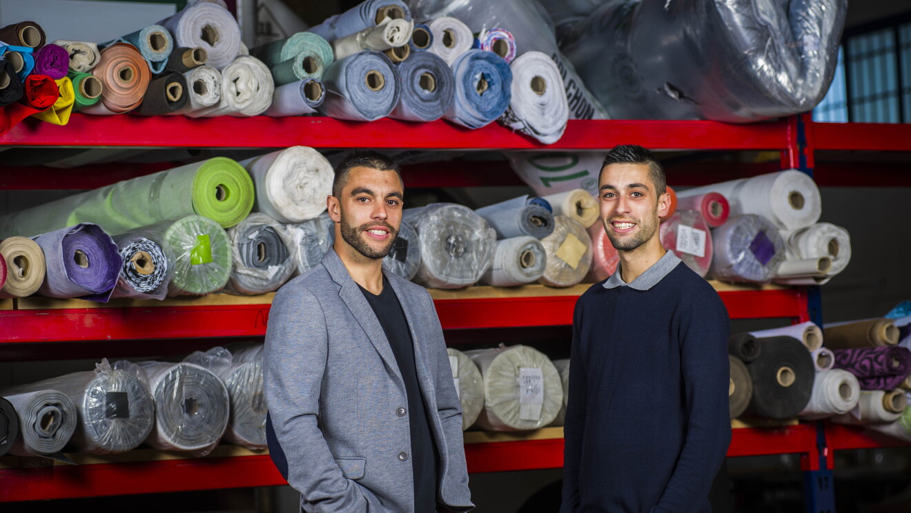 Los hermanos Javier y Daniel Mayorga con la marca SuenosZzz, los dos con las manos en los bolsillos, posan delante de las telas con las que tapizan sus productos. 