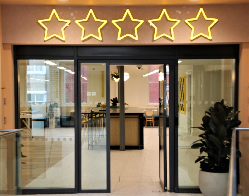 A view of the break area in the Amazon Manchester offices, this is viewed from outside of the open doors. Above the doors are neon lights depicting five stars. 