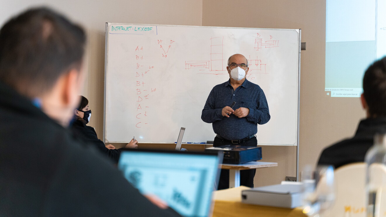 El programa Aprendizaje de Amazon tiene una parte teórica y otra práctica. En una aula y con una pizarra de rotulador y con una pantalla de ordenador proyectada a la pared, hay un profesor vestido con camisa azul, pantalones tejanos y un bolígrafo en la mano explicando teoría delante de la pizarra. Desenfocados se ven tres estudiantes sentados delante de sus ordenadores. 