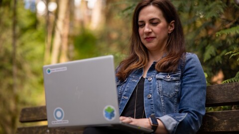 Ashely Rajagopal sits on a bench outside with vibrant greenery behind her. She is focused while she works on her laptop.
