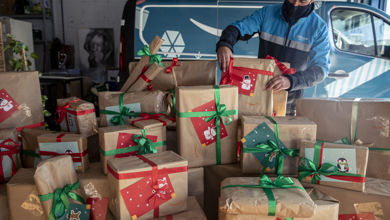 Centro de día para personas sin hogar Casablanca de Alcobendas. Una furgoneta de Amazon está dentro de un garaje. En segundo plano un conductor acabado de colocar una caja. El conductor va con una mascarilla negra y una chaqueta de Amazon azul claro, gris y azul marino. En primer plano paquetes de Amazon con lazos verdes y rojos y tarjetas regalo con motivos navideños.