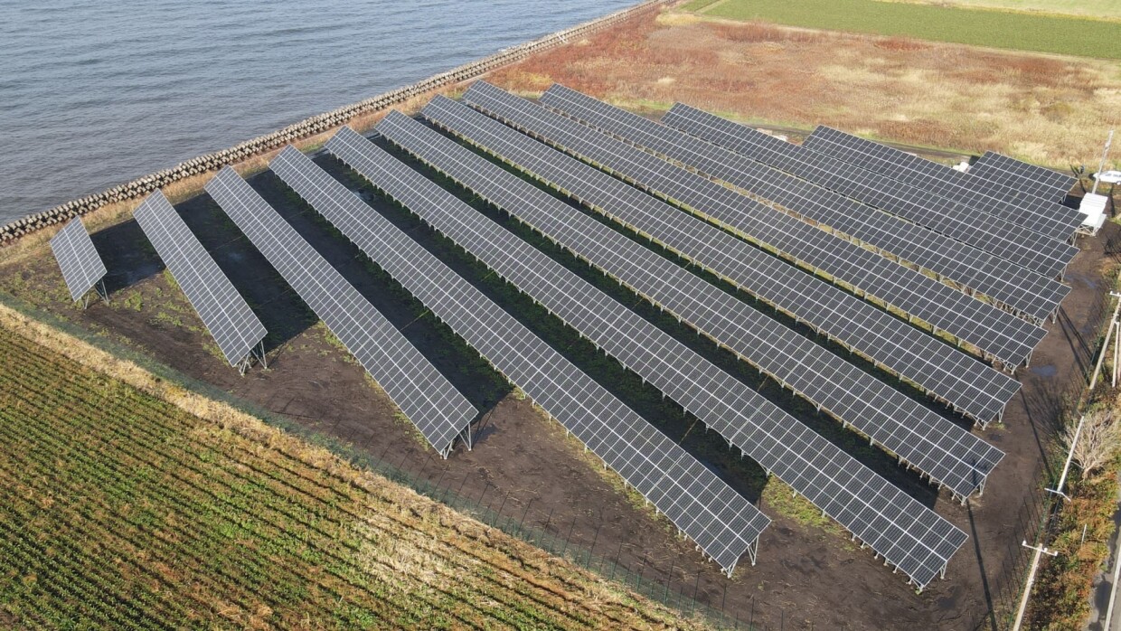 広大な畑に囲まれた土地に数十基の太陽光パネルが同じ方向を向いて設置されている。左上隅には水辺が見える