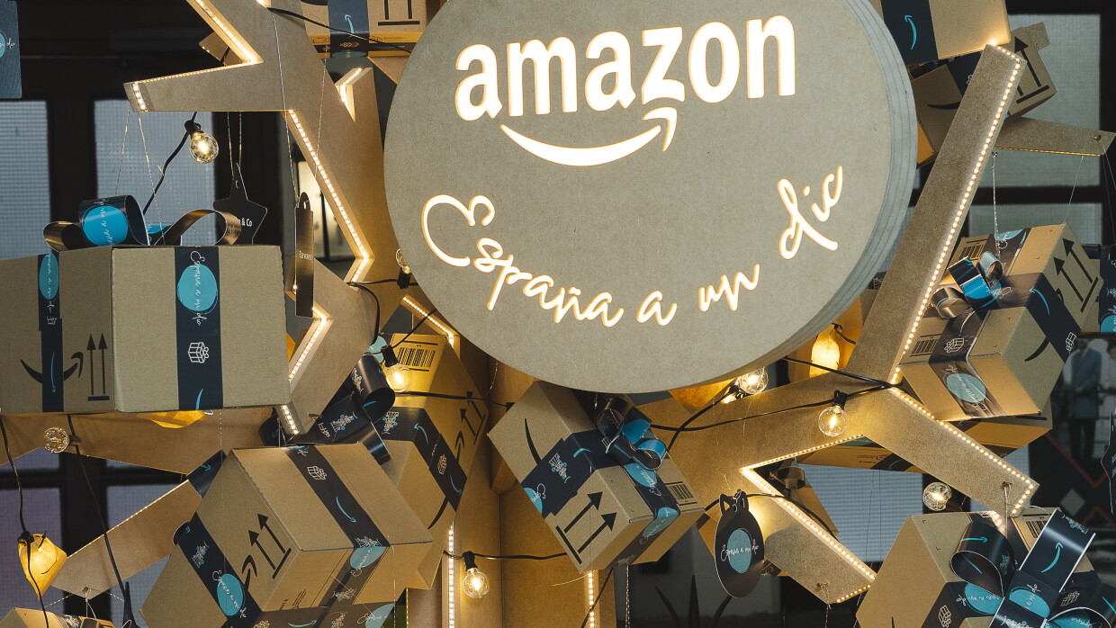 Árbol de navidad de cartón. Del Árbol cuelgan regalos que son cajas de Amazon y también figuras en forma de estrella y de bola de Navidad con el texto España a un clic. Hay también luces. Las cajas son de cartón marrón y los lazos, son el celo de Amazon de color negro y azul. En la cima del árbol hay una bola muy grande con el logo de Amazon y debajo el texto España a un clic. 