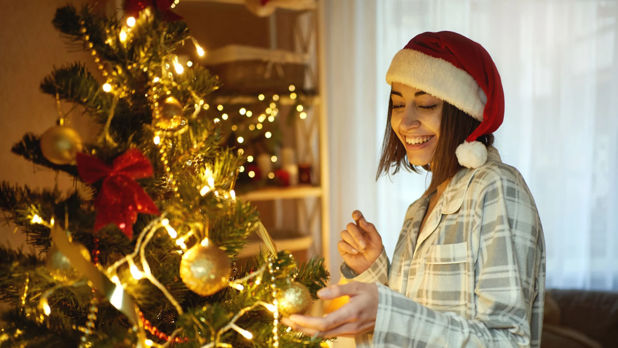 Mujer con un gorro de Papá Noel y vestida con pijama se encuentra decorando el árbol de Navidad.