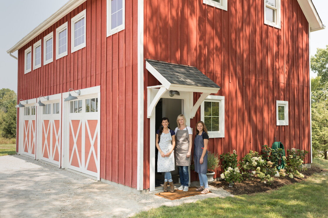 Three woman stand at the entrance of a tidy, red barn that has been converted into a workshop. All three women are wearing aprons.