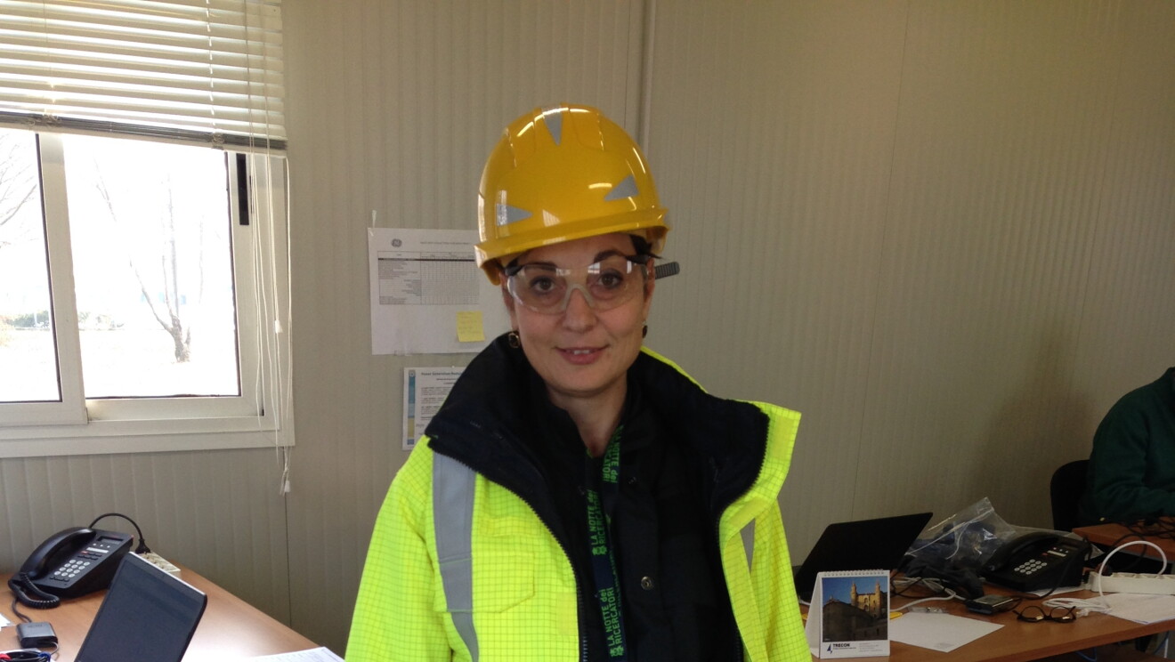 Donna in piedi in un ufficio in un centro operativo Amazon. Indossa un caschetto protettivo giallo
