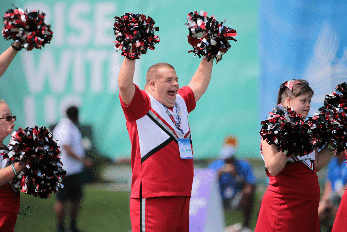 An image of a person with their arms up with a cheerleading uniform during the 2022 Special Olympics. There are other cheerleaders on the sides of the image holding their pom-poms.