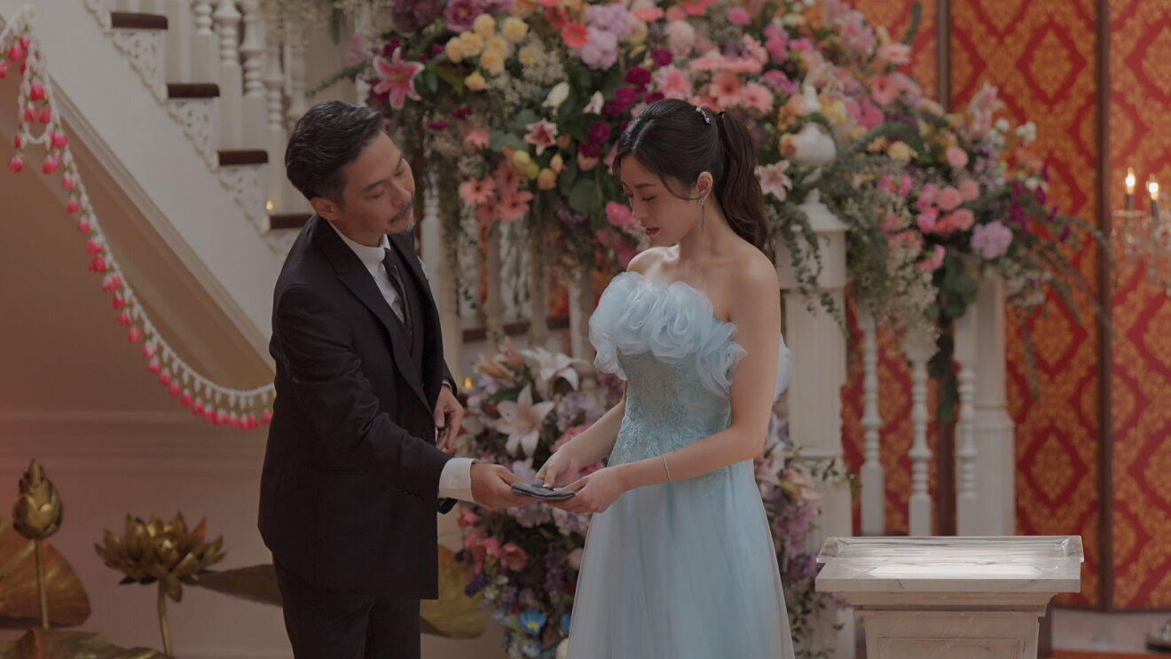 たくさんの花が飾られた豪華な室内に立つ2人の人物。左側の男性が、右側のドレス姿の女性に何かを手渡している。