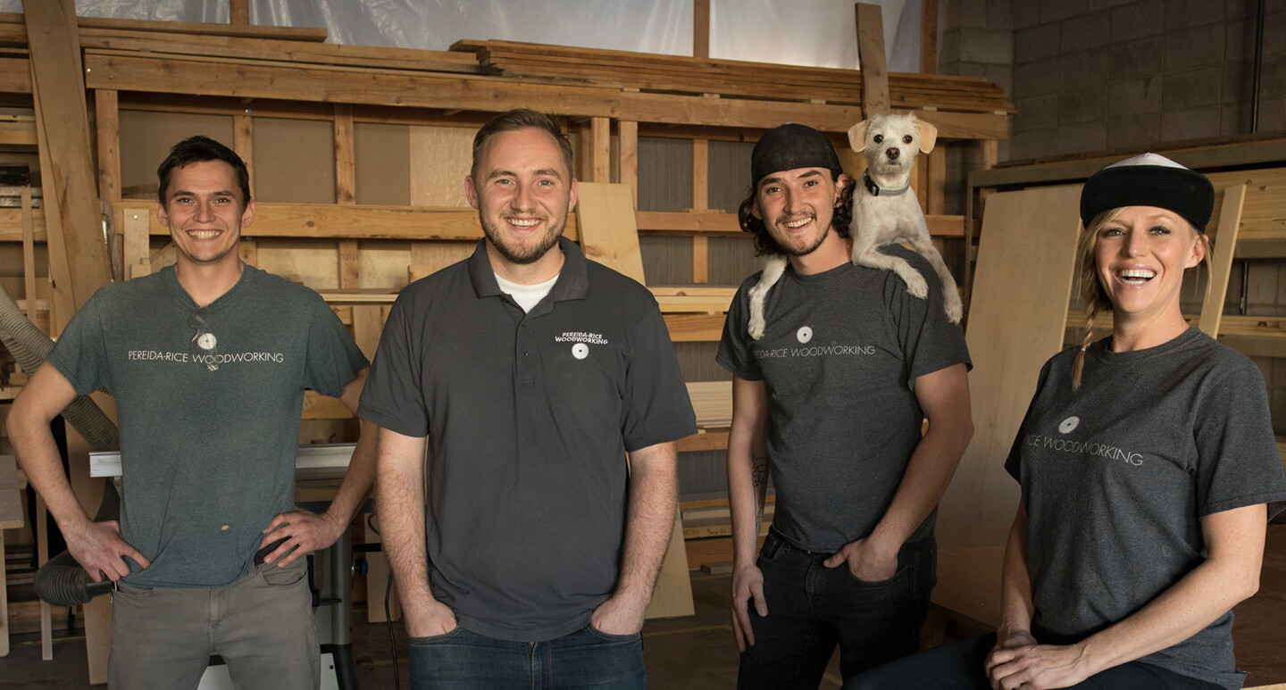 Three males and a female stand in front of a wall of wood. One of the men has a dog perched on his shoulders. These four individuals are siblings who run a small business named Pereida-Rice Woodworking, which sells on Amazon.