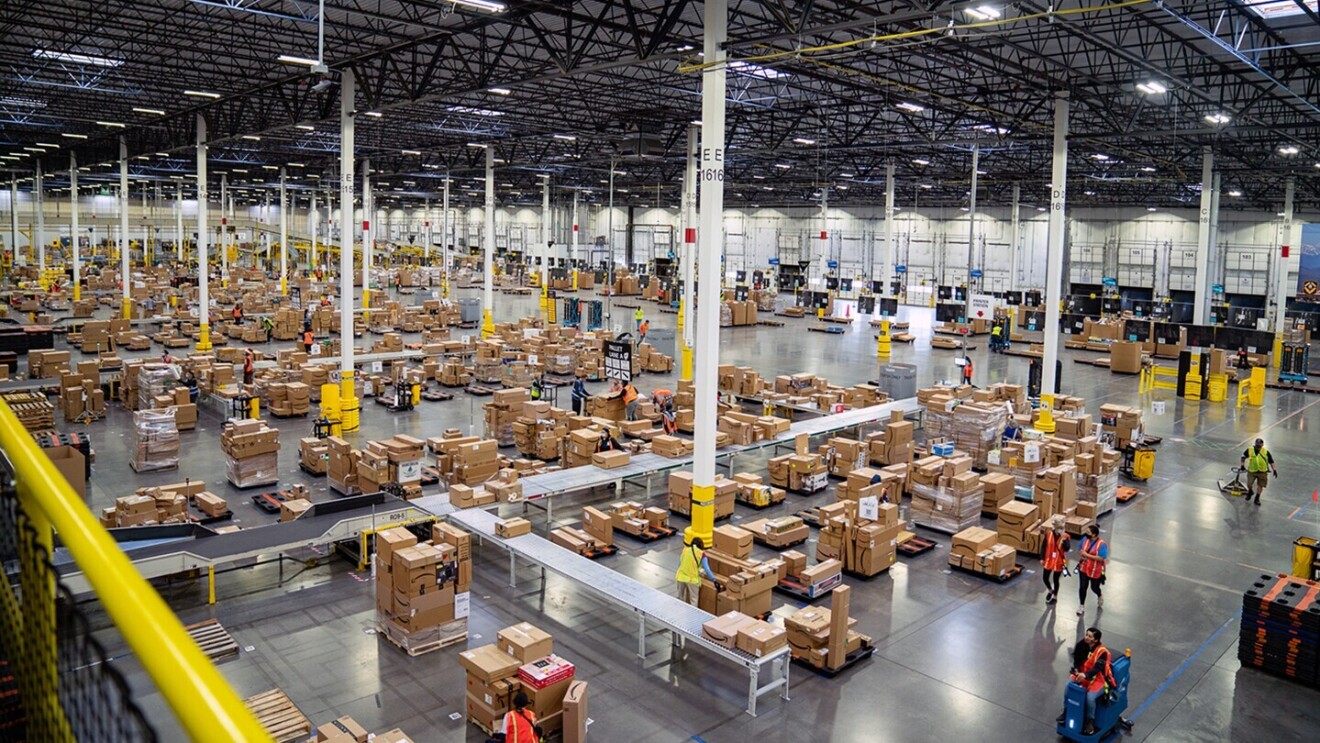 A view inside an Amazon fulfillment center (warehouse). Employees are seen moving about the space on foot, with hand trucks, and on a "mover" 