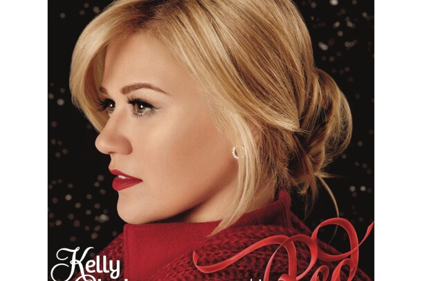 Kelly Clarkson wears her blonde hair tied behind her head, a red high-collared shirt with red scarf around her shoulders. Silver earrings, eye liner, and red lipstick completes her look. She looks to her right.
