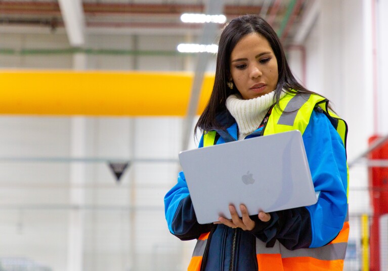 Ivette Duran, CEO de Cainaima Logistics, de pie con un ordenador entre las manos. De fondo está el centro logístico de color blanco y amarillo. Ella es morena y tiene el pelo largo y negro. Lleva un jersey de color blanco y de cuello alto, una chaqueta azul y un chaleco fluorescente.