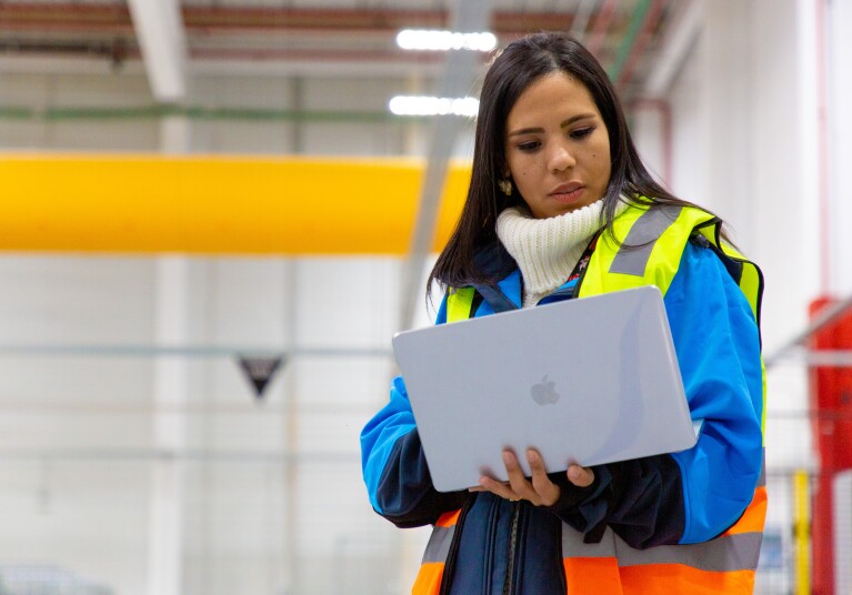 Ivette Duran, CEO de Cainaima Logistics, de pie con un ordenador entre las manos. De fondo está el centro logístico de color blanco y amarillo. Ella es morena y tiene el pelo largo y negro. Lleva un jersey de color blanco y de cuello alto, una chaqueta azul y un chaleco fluorescente. 