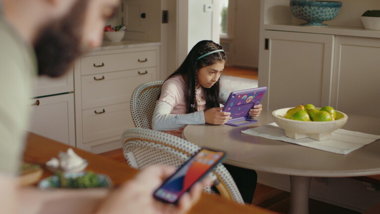 Girl sits at table viewing her tablet while dad is nearby on his smartphone.