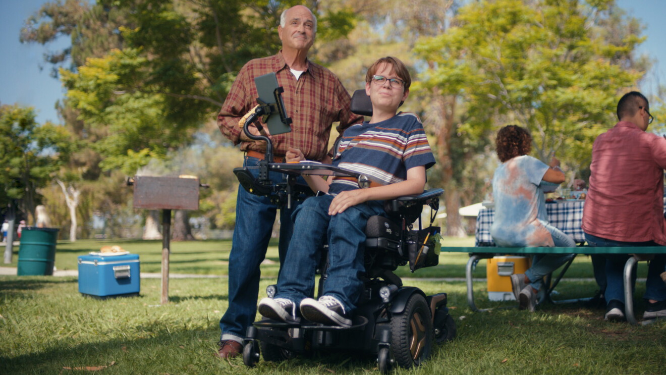 An image of a man and a boy in a park. The boy is in a wheelchair and the man is standing behind him.