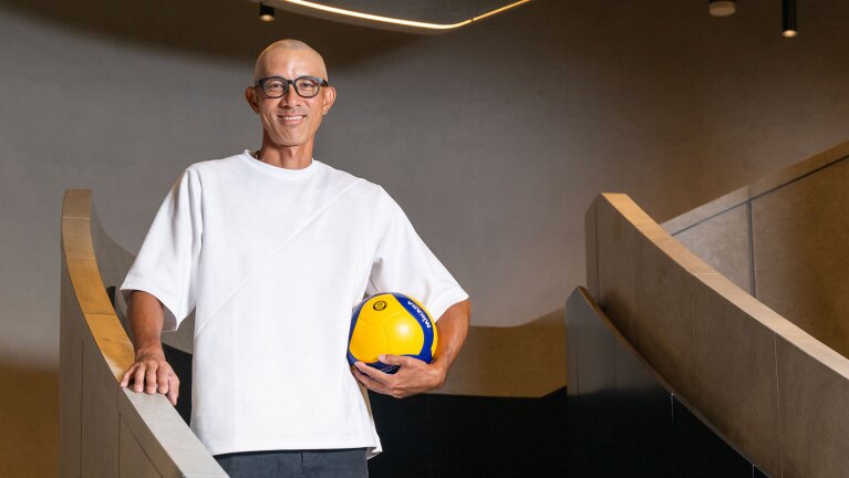 グレーの壁を背景に、木目調の手すりのある階段の途中に立つ人物。白い半袖Tシャツを着用し、黒縁の眼鏡をかけている。右手を木製の手すりに置き、左手で黄色と青のバレーボールを脇に抱え、こちらに笑顔を見せている