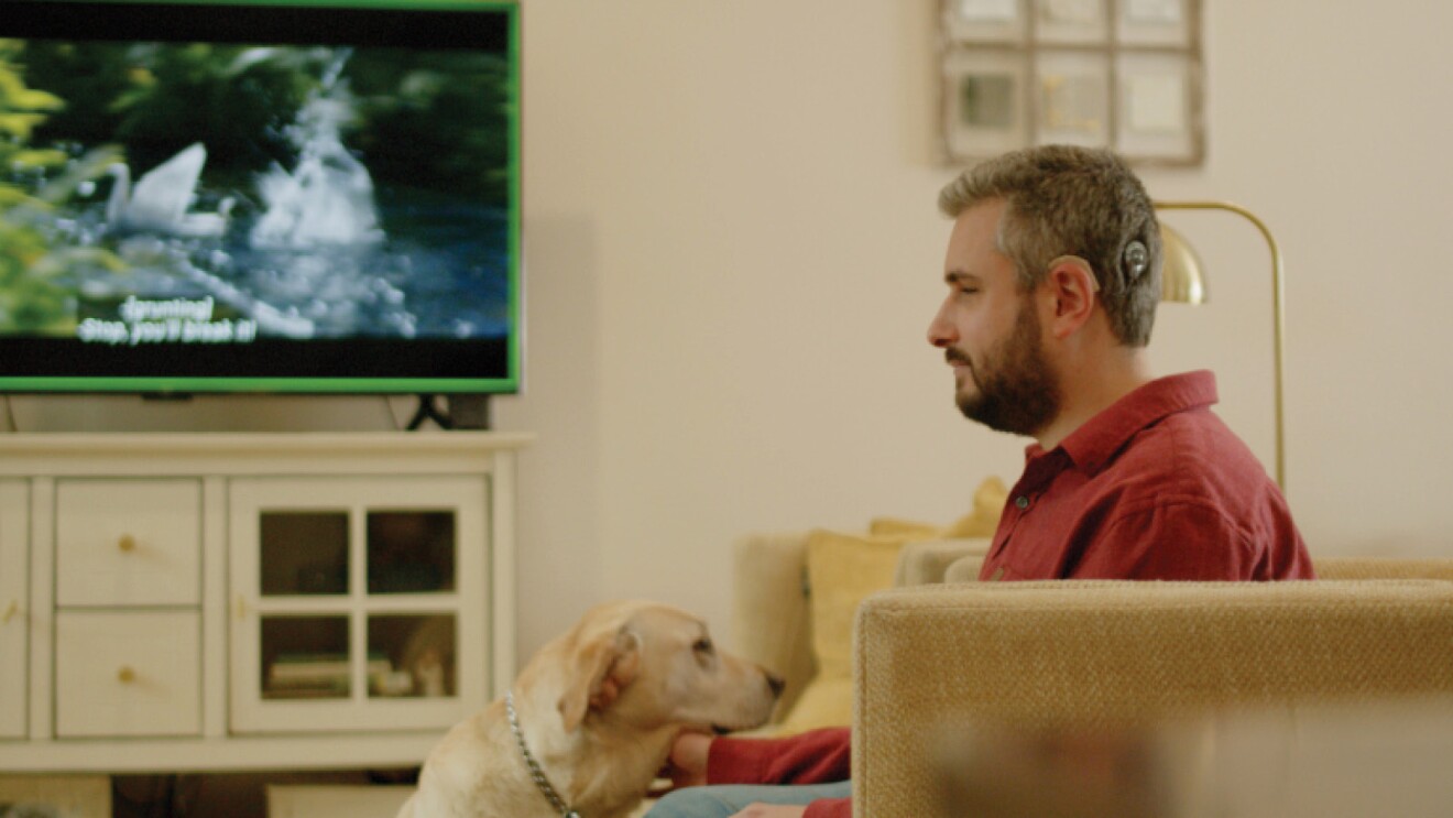 Photograph of Michael Forzano sitting on a couch in his living room petting his guide dog. In the background is a Fire TV with captions displayed.