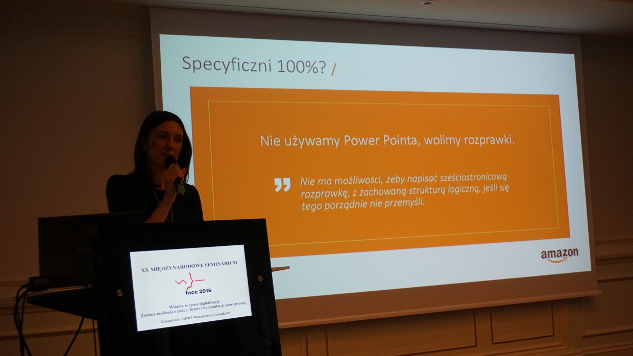 Prelegentka Amazon z mikrofonem, w tle slajd prezentacji z napisem: 'Nie używamy Power Pointa, wolimy rozprawki'