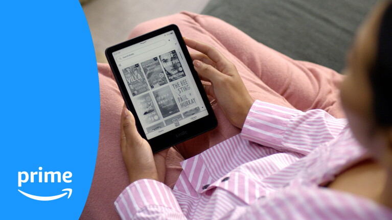 Person holding Kindle e-reader displaying library of book covers while relaxing on pink blanket