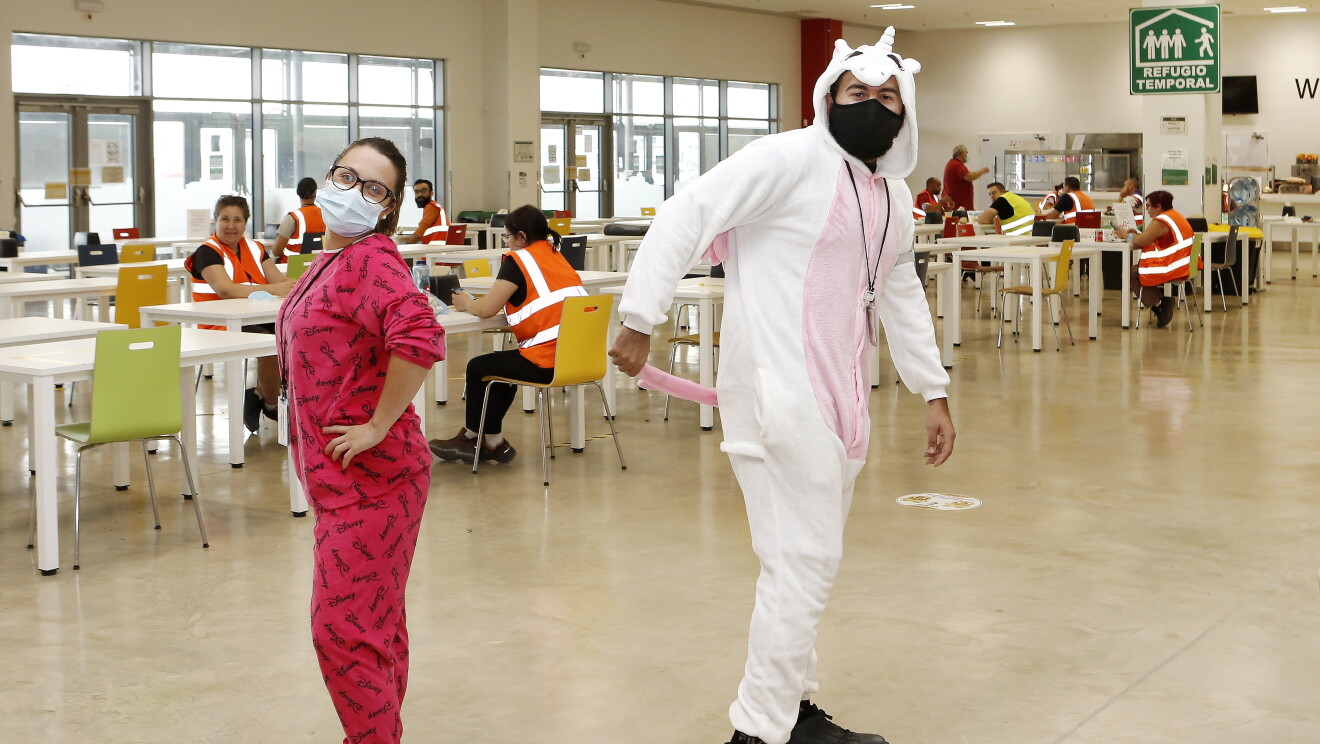 PJammin en MAD6 Illescas. En la cantina, de fondo asociados desayunando en mesas respetando la distancia de seguridad. Dos asociados dándose al espalda y en pijama. Los dos con mascarilla. Una chica va con un pijama rosa, tiene las manos en la cintura, gafas y mascarilla y se da la espalda con  su compañero vestido de unicornio con un pijama que tiene gorro. Se agarra la cola con una mano. Lleva mascarilla negra. 