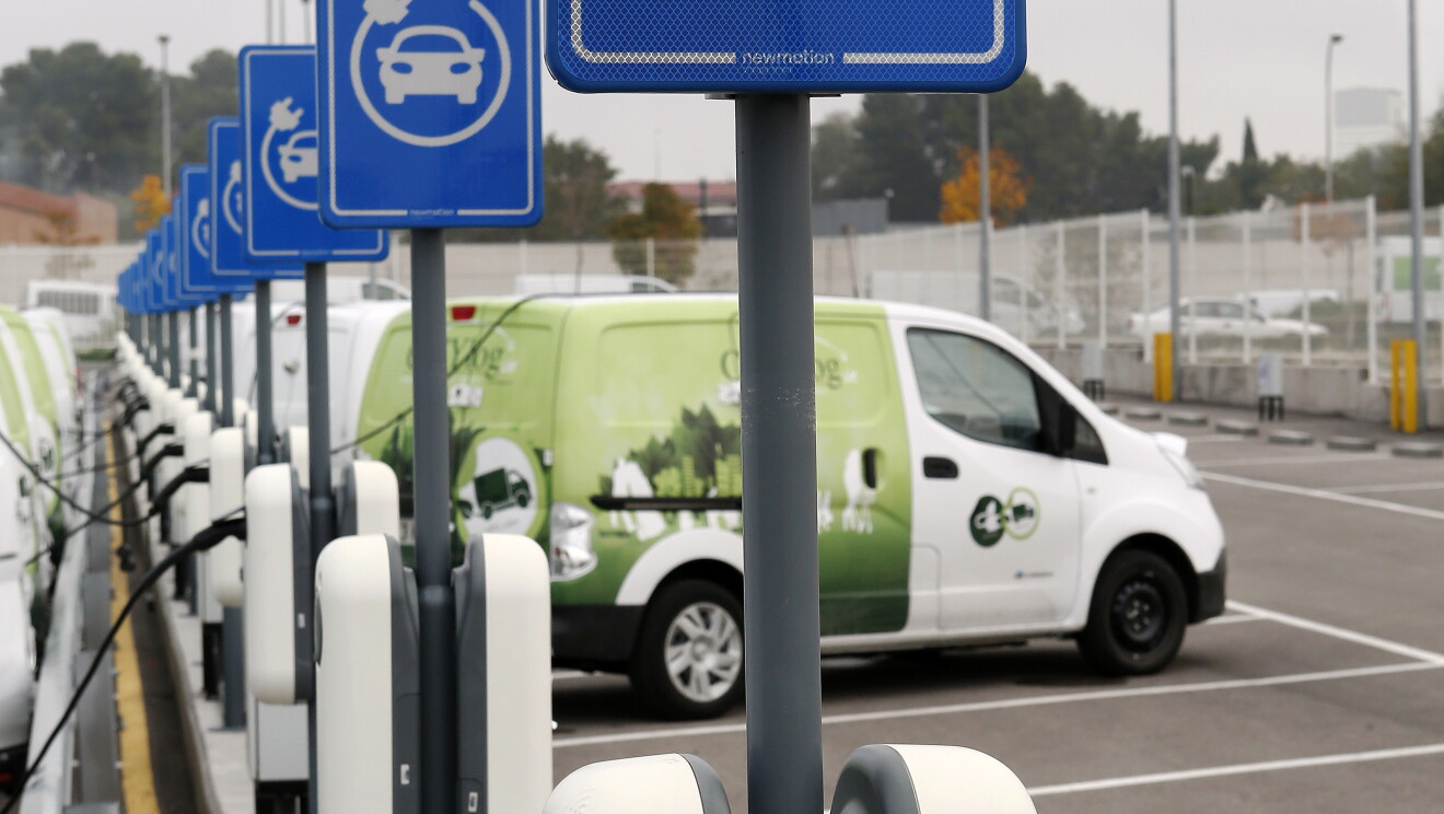 Cargado eléctrico en primer plano. De colo blanco con un cable conectado a la corriente. Se ve un seguido de cargadores hasta el fondo de la foto y unas cuatro furgonetas cargando  batería. Es el aparcamiento del centro logístico de Madrid, 