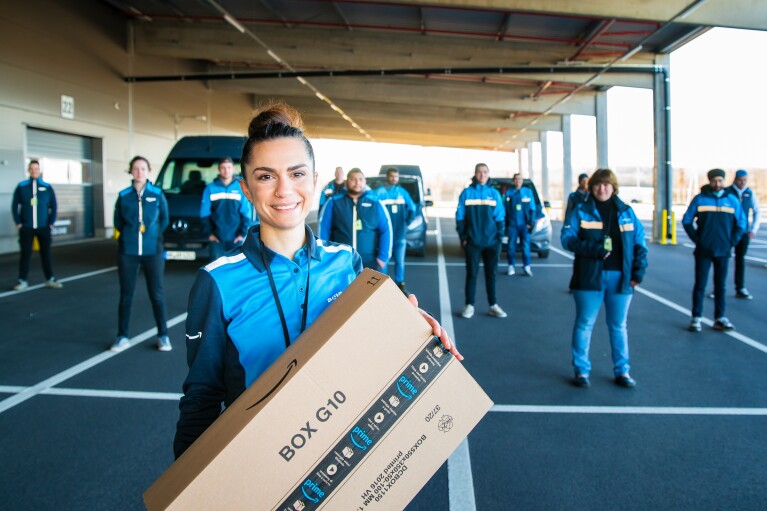 Eine zierliche junge Frau steht im Vordergrund mit einem Amazon Paket, hinter ihr sind 30 Mitarbeiter:innen verteilt auf einem Parkplatz zu sehen. Alle tragen Arbeitskleidung im Amazon Fahrer:innen Branding.