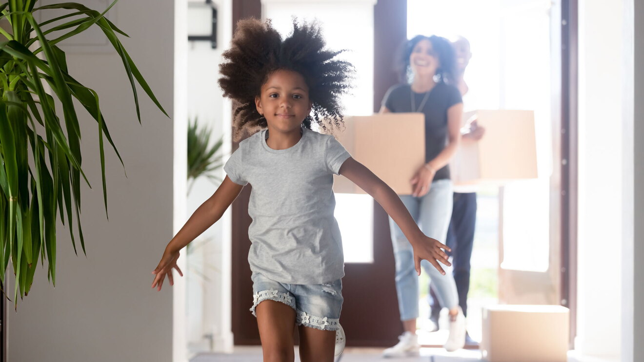 A little girl runs into a house.  Adults carrying moving boxes into the home are out of focus behind her.