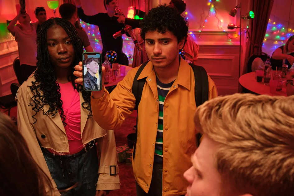 A man and a woman stand in the middle of a party holding a mobile phone 