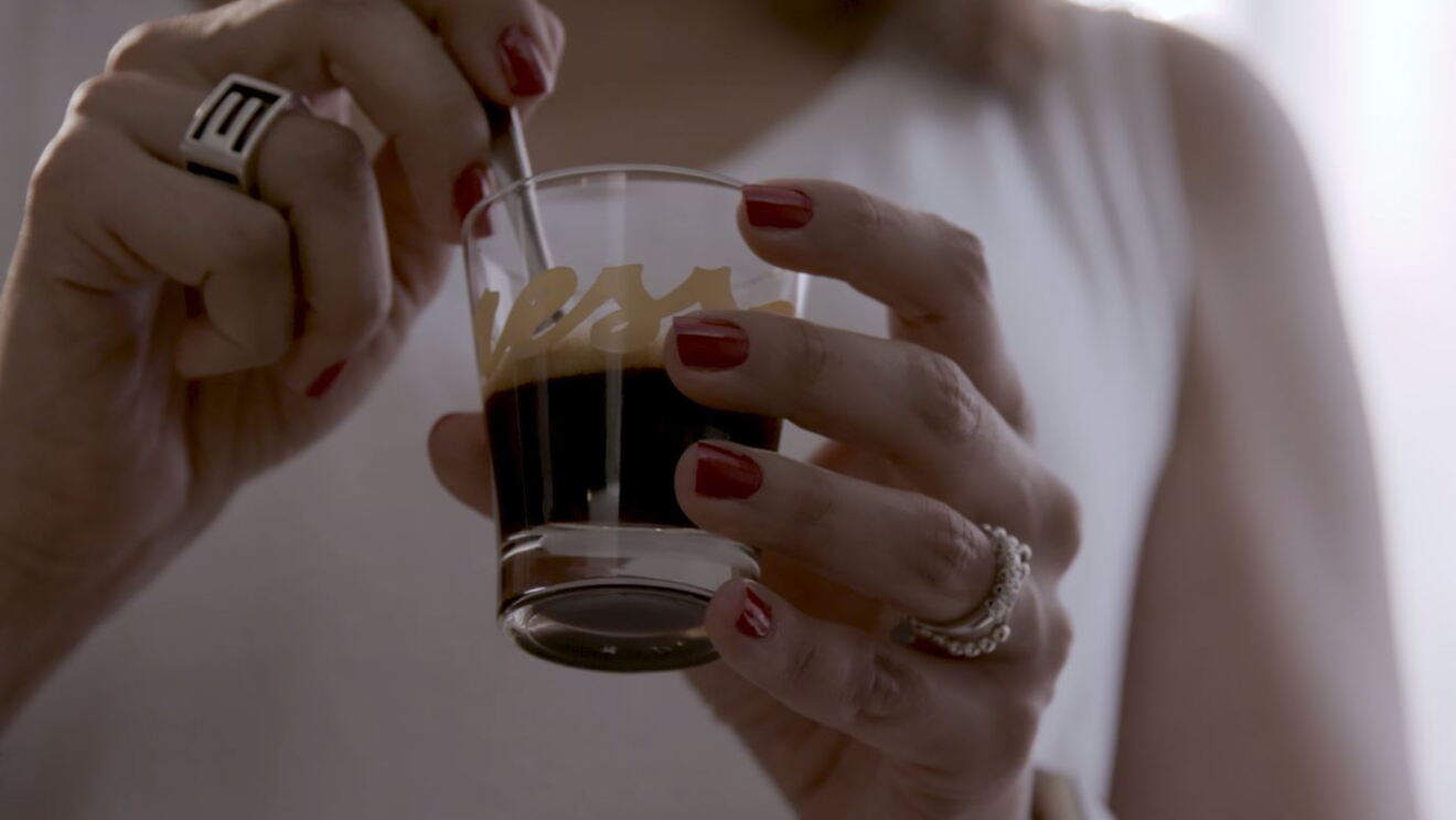 Primo piano di due mani femminili che reggono una tazzina di caffè in vetro. Sulla tazzina si intravede il logo 'Yespresso'. La donna, di cui non si vede il volto, indossa una canotta bianca, ha due anelli su entrambi gli anulari e lo smalto rosso. La sua mano sinistra regge la tazzina, mentre con la destra gira il caffè con un cucchiaino. 