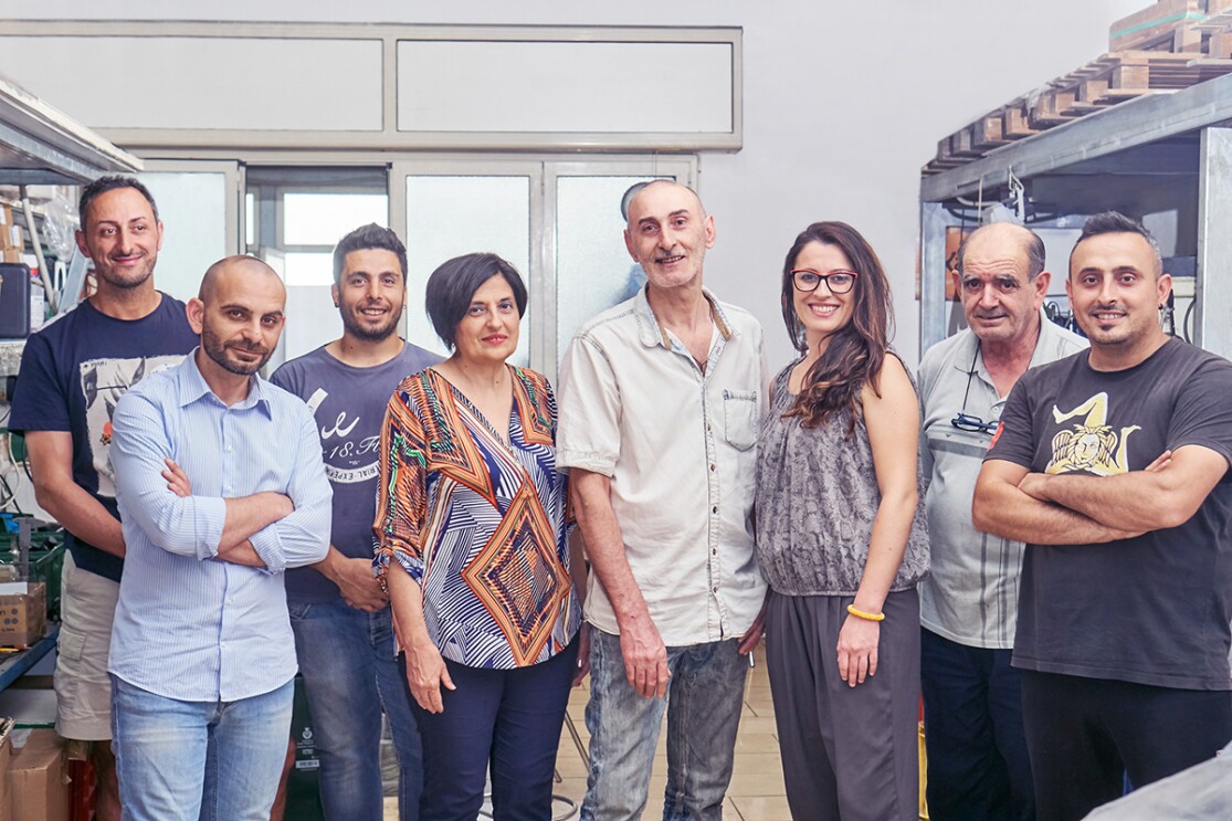 Foto del team di Kamira, formato da 8 persone in piedi, 6 uomini e 2 donne, tutti sorridenti, inclusi il fondatore Nino Santoro e la sua famiglia.