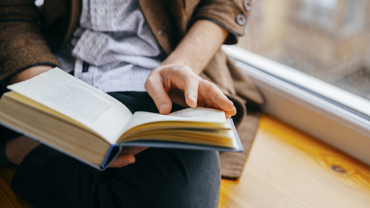 A person sits on a window ledge, while reading a book