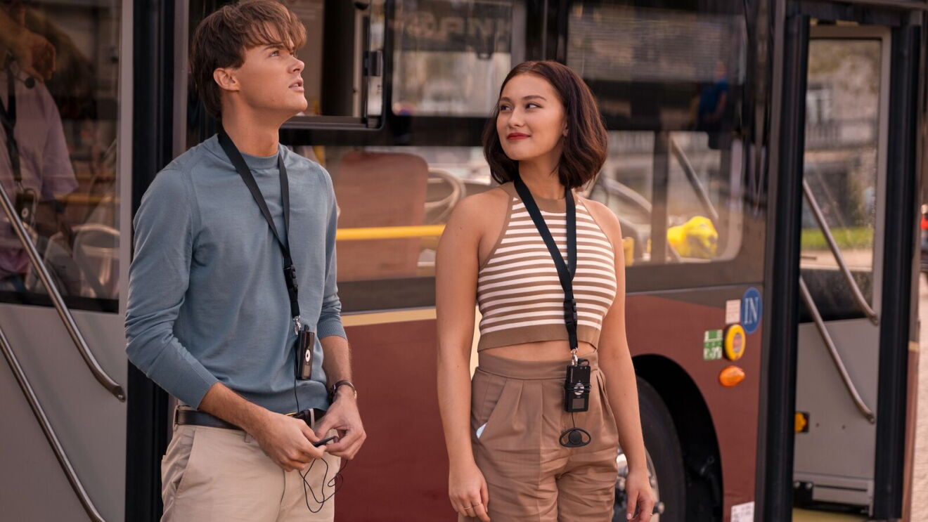 A scene from The Summer I Turned Pretty with two people with lanyards standing in front of a bus, dressed casually for summer
